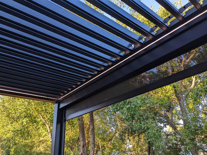 The dark motorized louvered pergola features partially open louvers that cast slender linear shadows. Its structure is made of dark pillars and beams, with warm yellow LED strips subtly glowing along the edges. A dark shade screen on one side is partially lowered for added sun protection.