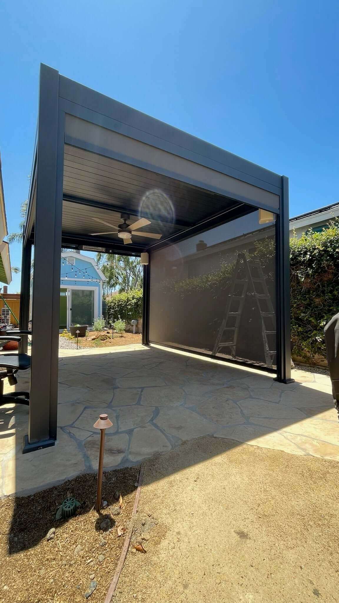 This motorized louvered pergola features a partially open top, revealing its sleek slatted design. The deep gray aluminum frame has clean, modern lines. A light-colored outdoor ceiling fan is installed underneath. Deep gray sunshade blinds on the sides are partially lowered, offering shade and some privacy. The blinds are semi-translucent, blocking direct sunlight while allowing soft, filtered light through.