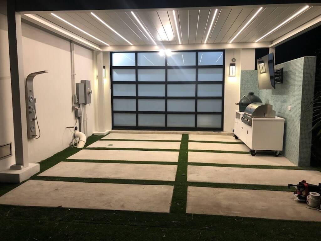 This area is covered by a white Bon Pergola, featuring a roof with adjustable white louvers and integrated LED strip lighting that illuminates the space below. The ground beneath is finished with light gray flooring, divided into sections by vibrant green artificial turf, adding a touch of freshness and texture. On one side of the pergola, a black-framed sliding glass door connects the space, creating a seamless transition. The overall vibe is modern, comfortable, and fully functional.