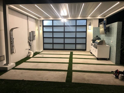This area is covered by a white Bon Pergola, featuring a roof with adjustable white louvers and integrated LED strip lighting that illuminates the space below. The ground beneath is finished with light gray flooring, divided into sections by vibrant green artificial turf, adding a touch of freshness and texture. On one side of the pergola, a black-framed sliding glass door connects the space, creating a seamless transition. The overall vibe is modern, comfortable, and fully functional.