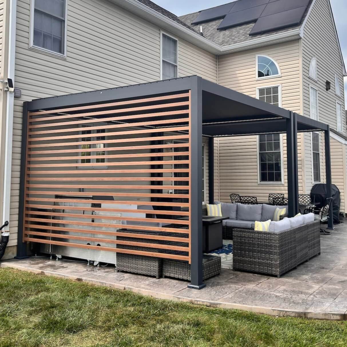 The main focus of the photo is a black modern pergola with motorized louvers on the top. One side features horizontally arranged wood-grain louver panels, providing both privacy and a stylish look. The entire structure is sleek, made up of black columns and beams.