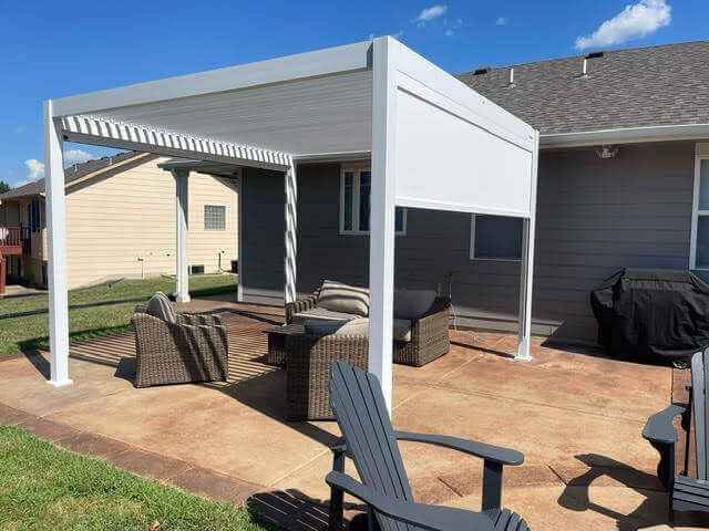 The BON PERGOLA features a partially open louvered top, casting striped shadows below. The structure is made of sturdy white posts and beams. White motorized blinds are installed on the sides, with part of the blinds lowered to provide shade and privacy. The blinds appear thick, effectively blocking sunlight. With the adjustable motorized blinds, the BON PERGOLA allows for tailored shading based on weather and needs, creating a comfortable outdoor space.