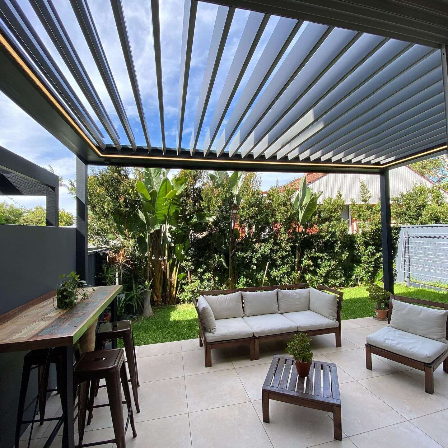 A dark gray Bon Pergola electric louvered pergola features bold louvers with a warm yellow LED strip embedded along the edge—visible even in daylight. Its structure is built with thick, modern posts and beams in matching deep gray. A potted plant sits atop the coffee table below, adding a cozy detail. Surrounded by vibrant greenery, including banana trees and other tropical plants, the space feels alive and lush—perfect for relaxing in a stylish, nature-filled outdoor setting.